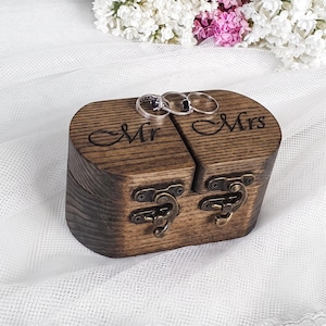 May include: A dark brown wooden ring box with two compartments, each secured by a bronze-colored metal latch. The lid features the engraved text Mr and Mrs. Three silver-toned metal rings are resting on the top surface of the wooden box.