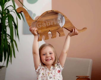 May include: A wooden dinosaur-shaped money box featuring a clear acrylic front panel. The name Gabriel is engraved in the centre of the transparent window. The box is constructed from light brown wood with a visible grain and secured with small screws.