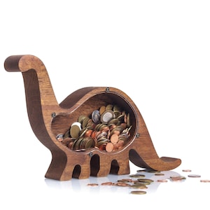 Op de afbeelding: Een dinosaurusvormige houten spaarpot met een donkerbruine afwerking en een doorzichtig acrylpaneel aan de zijkant. De binnenkant is gevuld met diverse metalen munten en er liggen enkele losse munten verspreid voor het object.