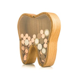 May include: A wooden tooth-shaped bank filled with various coins. The bank is made of light-colored wood with a clear plastic front, allowing a view of the coins inside. The coins are a mix of copper and silver colors.