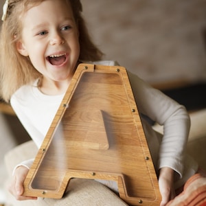 A young girl is sitting on a couch, holding a wooden letter-shaped piggy bank and smiling.