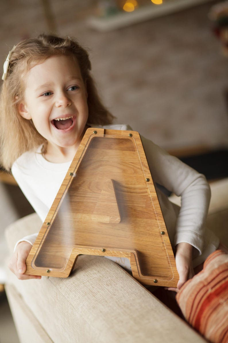 A young girl is sitting on a couch, holding a wooden letter-shaped piggy bank and smiling.