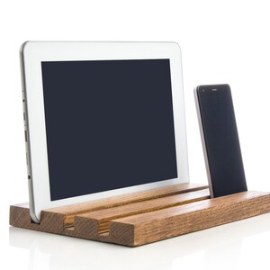 May include: A wooden stand with two slots for holding a tablet and a smartphone. The stand is made of dark brown wood and has a natural finish. The tablet and smartphone are both black and have blank screens.