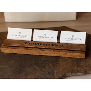 May include: A rectangular wooden business card holder with a dark brown wood grain finish. The front features the text WOODPRESENTS engraved in black. Three white business cards with the logo and text WOODPRESENTS HANDCRAFT WORKSHOP are inserted.