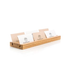 May include: A rectangular light brown wooden stand with a horizontal groove holding four white rectangular cards. Each card features a black graphic of a gift box and the text WoodPresents printed in a clear, dark font on the front surface.