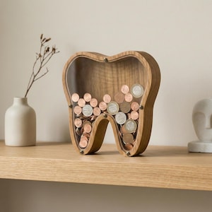 May include: A wooden tooth-shaped coin bank filled with various coins. The bank has a clear acrylic front, allowing visibility of the coins inside. The bank is made of light brown wood and sits on a wooden shelf.