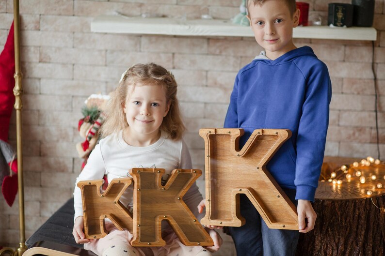 A young girl and boy are holding wooden letters, K and K, in front of a brick wall.