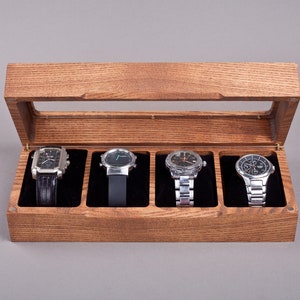 May include: A wooden watch box with a glass top, containing four watches with silver bands and black faces.