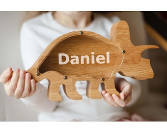 May include: A light brown wooden dinosaur-shaped money box with a clear acrylic front panel. The name Daniel is printed in white letters on the centre of the acrylic. The wooden frame is secured with small metal screws around the perimeter.
