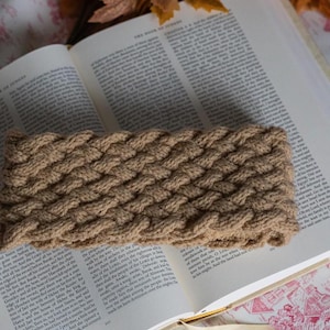 May include: A brown knitted headband with a braided pattern. The headband is lying on a book with text.