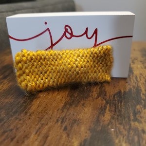 May include: A small, yellow knitted rectangle with a textured surface. The rectangle is sitting on a wooden surface in front of a white sign with the word "joy" written in red.