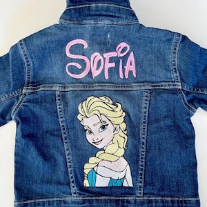 May include: A blue denim jacket with the name "Sofia" embroidered in pink on the back. The jacket features an embroidered image of a blonde princess in a blue and white dress.