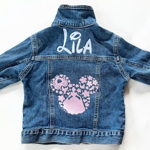 May include: A blue denim jacket with the name "Lila" in white script across the upper back. The lower back features a pink embroidered design of a princess in a dress, surrounded by hearts and flowers.