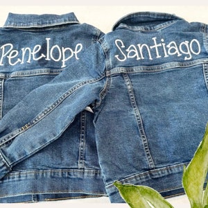 May include: Two denim jackets with white embroidered names on the back. The jacket on the left says "Penelope" and the jacket on the right says "Santiago".
