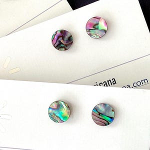 May include: Stud earrings with iridescent abalone shells, displayed on white cards. The earrings have a circular shape and showcase a range of colors, including pink, green, and blue. The cards feature the text "Artesana Americana" and "handmade".