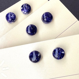 May include: Six round, dark blue stud earrings with white speckles are displayed on cream-colored earring cards. The earrings are arranged in two rows of three. The cards have the text "eclectic | handmade" and the logo and name "Artesana Americana".