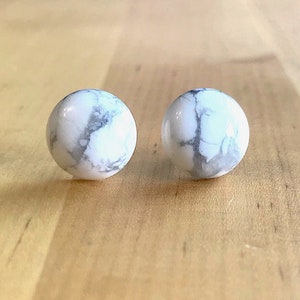 May include: Two round, white marble earrings with gray veining. The earrings are set against a light brown wooden surface. These earrings are a classic accessory.
