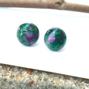 May include: A pair of green and pink gemstone stud earrings. The earrings are round and have a speckled appearance.