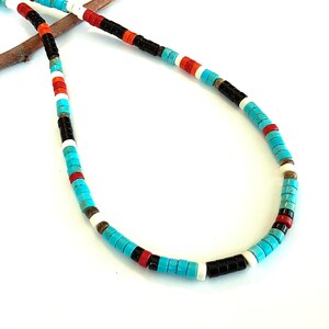 May include: A necklace made of turquoise, black, red, white, and brown beads. The beads are small and cylindrical, strung together in a repeating colour pattern. The necklace is displayed on a small, brown twig. The necklace is approximately 45 cm in length.