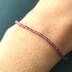 May include: A delicate bracelet made with small, round, pink-purple beads. The bracelet is shown on a person's wrist.