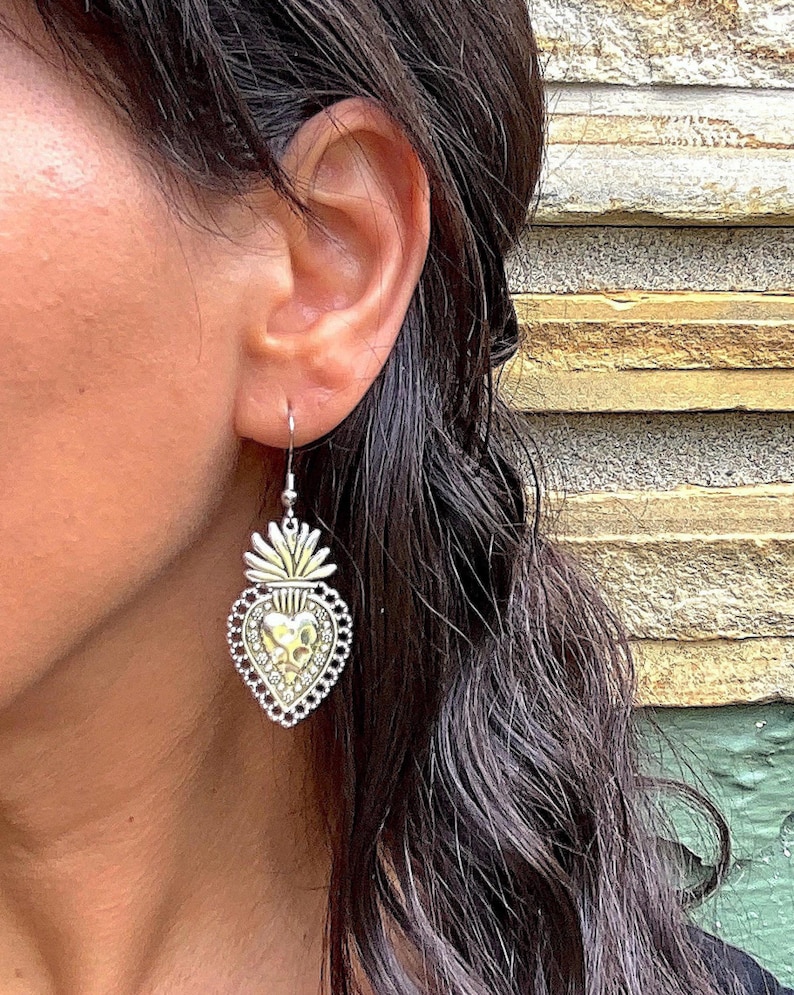 May include: Silver heart-shaped earrings with a gold-toned design and a detailed floral pattern. The earrings are hanging from a person's ear.