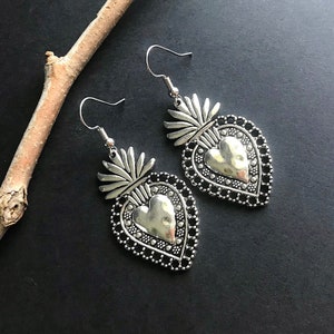 May include: A pair of silver earrings with a heart-shaped design. Each earring features a detailed heart with a raised center and a decorative border. The hearts are attached to a small, ornate floral design.