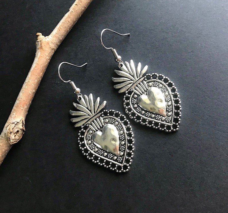 May include: A pair of silver earrings with a heart-shaped design. Each earring features a detailed heart with a raised center and a decorative border. The hearts are attached to a small, ornate floral design.