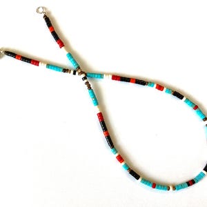 May include: A beaded necklace featuring turquoise, red, black, white, and brown beads. The necklace has a simple design with a clasp closure. The beads are small and evenly spaced, creating a colourful and eye-catching accessory.