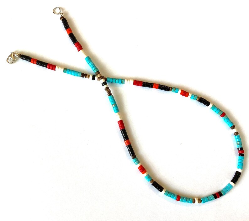 May include: A beaded necklace featuring turquoise, red, black, white, and brown beads. The necklace has a simple design with a clasp closure. The beads are small and evenly spaced, creating a colourful and eye-catching accessory.