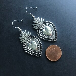 May include: Silver tone earrings with a heart-shaped design and a detailed filigree border. The heart has a textured surface and is surrounded by a decorative border. The earrings are approximately the size of a US penny.