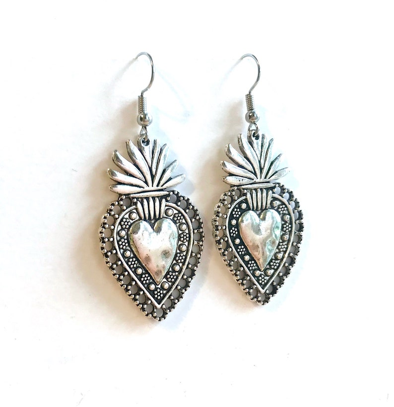 May include: Silver heart-shaped earrings with a detailed design and a textured heart in the center. The earrings have a hook closure.