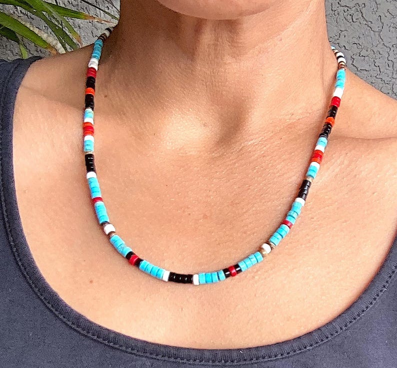 May include: A multi-coloured beaded necklace with turquoise, red, black, white, and orange beads. The necklace is made of small, round beads strung together, creating a vibrant accessory. The necklace is worn around the neck.