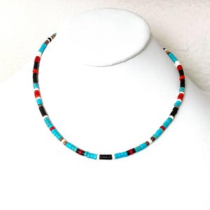 May include: A vibrant beaded necklace featuring turquoise, red, black, and white beads. The necklace is presented on a white mannequin bust, highlighting its design and colour arrangement. The beads are small and evenly spaced.