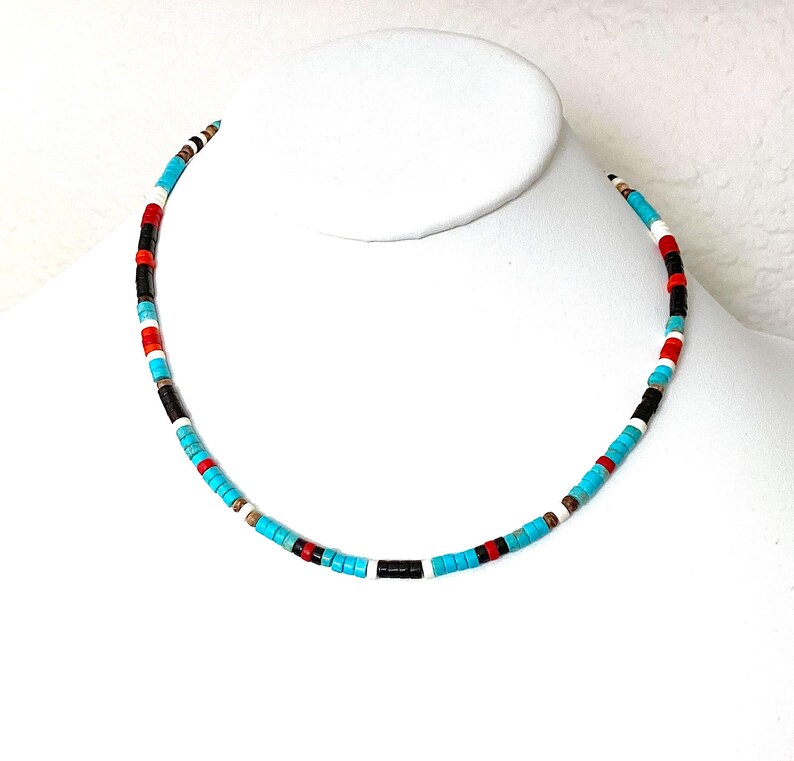 May include: A vibrant beaded necklace featuring turquoise, red, black, and white beads. The necklace is presented on a white mannequin bust, highlighting its design and colour arrangement. The beads are small and evenly spaced.