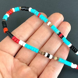 May include: A vibrant beaded necklace featuring turquoise, red, white, and black beads. The necklace is strung on a thin cord. The beads are arranged in a repeating pattern. The background is dark.