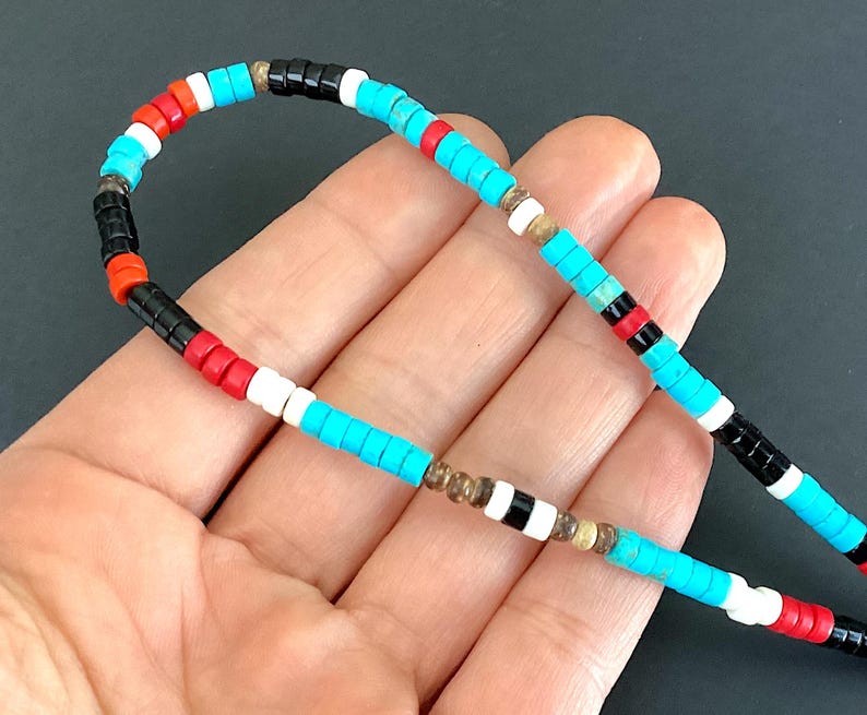 May include: A vibrant beaded necklace featuring turquoise, red, white, and black beads. The necklace is strung on a thin cord. The beads are arranged in a repeating pattern. The background is dark.