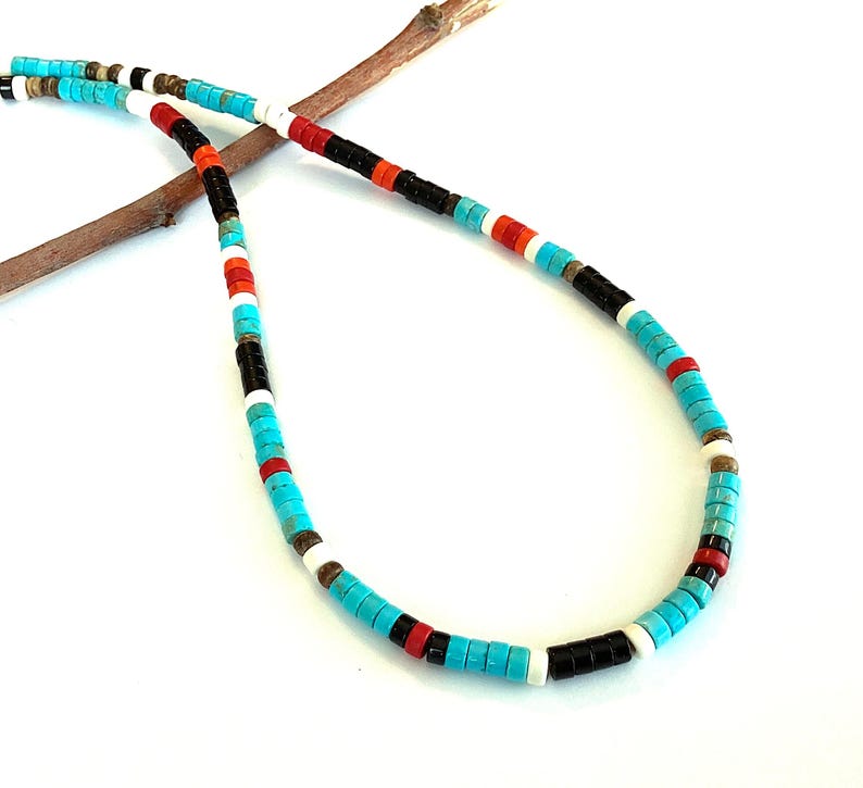 May include: A necklace made of turquoise, black, red, white, and brown beads. The beads are small and cylindrical, strung together in a repeating colour pattern. The necklace is displayed on a small, brown twig. The necklace is approximately 45 cm in length.