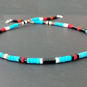 May include: A necklace made of small, cylindrical beads in turquoise, black, red, and white. The necklace is strung on a silver-coloured chain. The beads are displayed against a dark background.
