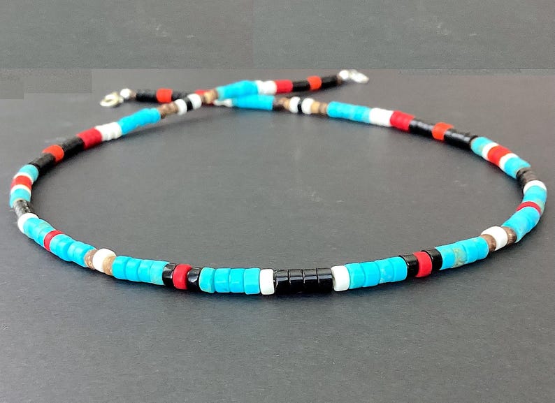 May include: A necklace made of small, cylindrical beads in turquoise, black, red, and white. The necklace is strung on a silver-coloured chain. The beads are displayed against a dark background.