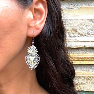 May include: Silver heart-shaped earrings with a detailed design and a small, gold-colored heart in the center. The earrings are hanging from a person's ear.