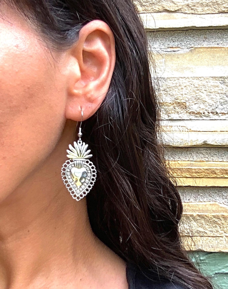 May include: Silver heart-shaped earrings with a detailed design and a small, gold-colored heart in the center. The earrings are hanging from a person's ear.