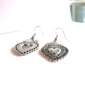 May include: A pair of silver heart-shaped earrings with a detailed design. The earrings have a textured surface and a small loop at the top for hanging.