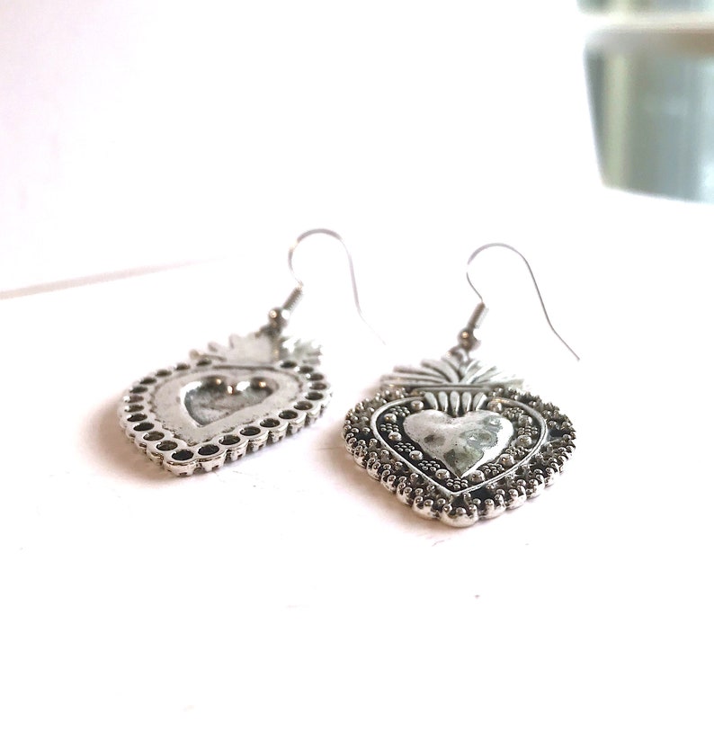 May include: A pair of silver heart-shaped earrings with a detailed design. The earrings have a textured surface and a small loop at the top for hanging.