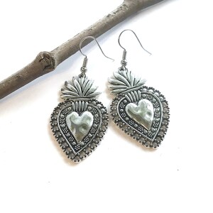 May include: Silver heart-shaped earrings with a textured heart design and a crown on top. The earrings are hanging from a branch.