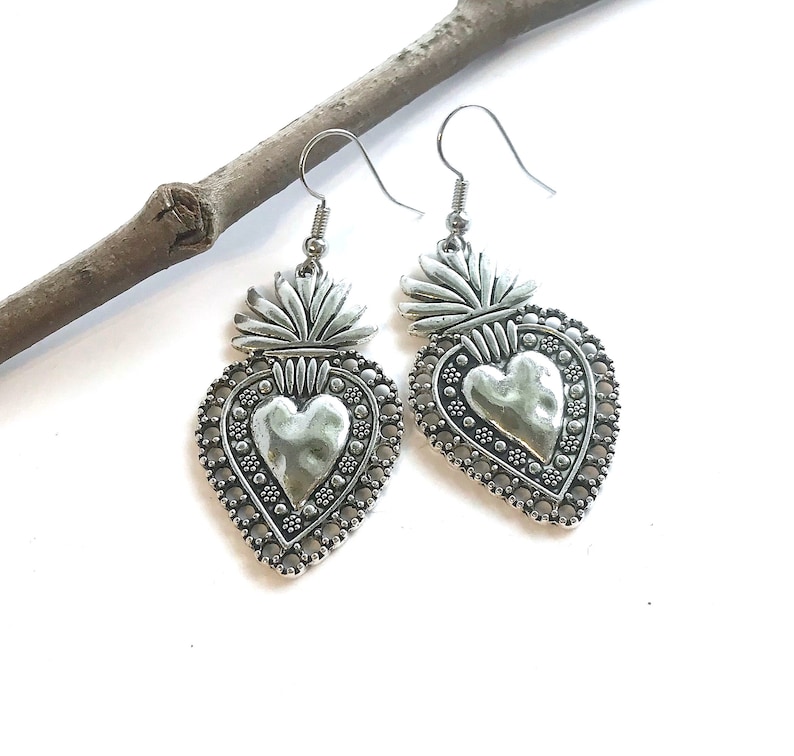 May include: Silver heart-shaped earrings with a textured heart design and a crown on top. The earrings are hanging from a branch.