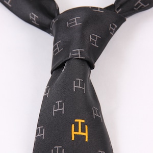 Puede incluir: Una corbata negra con un patrón repetido de letras "H" grises. La corbata está anudada, con una sola "H" amarilla en la parte inferior. La corbata está hecha de un material liso, posiblemente seda.