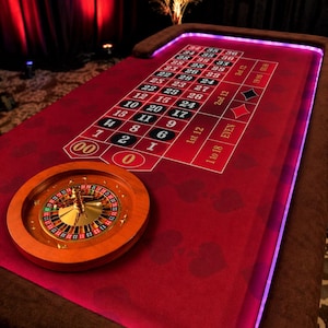 May include: A casino roulette table with a red felt surface and a wooden roulette wheel. The table has numbers and betting areas, with a brown border and purple lighting. The roulette wheel is made of wood and metal.