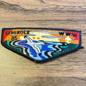 May include: A triangular embroidered patch featuring a sunset landscape with a white bird flying over blue water. The text SEMINOLE 85 is stitched in black on the left, and WWW is stitched in black on the right, all bordered by a thick black edge.