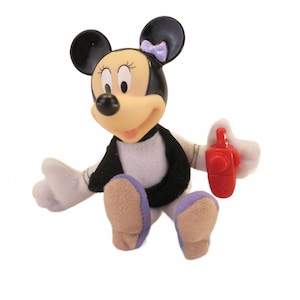 May include: A Minnie Mouse figurine with a black and white outfit, purple shoes, and a purple bow. The figurine is holding a red object. The character has a tan face and black ears.