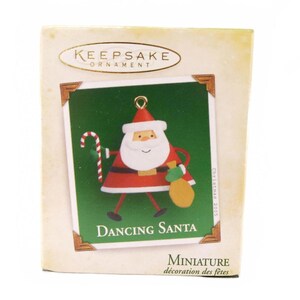 May include: A miniature Christmas ornament in a box. The ornament features a cartoon Santa Claus with a red suit, white beard, and a candy cane. The text "Dancing Santa" is below the image. The box reads "Keepsake Ornament".